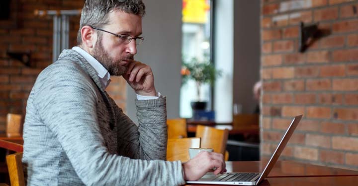 A middle-aged man with glasses and a beard sits at a table in a cafe, looking intently at a laptop. He is wearing a grey cardigan over a white shirt. The background shows a brick wall and blurred cafe seating. Text overlays the image, stating: "We're here to help! Let's learn more about how to name a trustee and use a deed of trust."