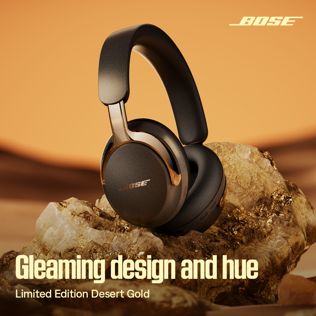 A close-up, studio-style shot of a pair of black Bose headphones with gold accents is displayed against a warm, gradient orange background. The headphones are resting on a textured surface resembling rocks with gold flecks. Overlayed text in a bold, yellow font reads "Gleaming design and hue" and "Limited Edition Desert Gold". The Bose logo is visible in the top right corner.