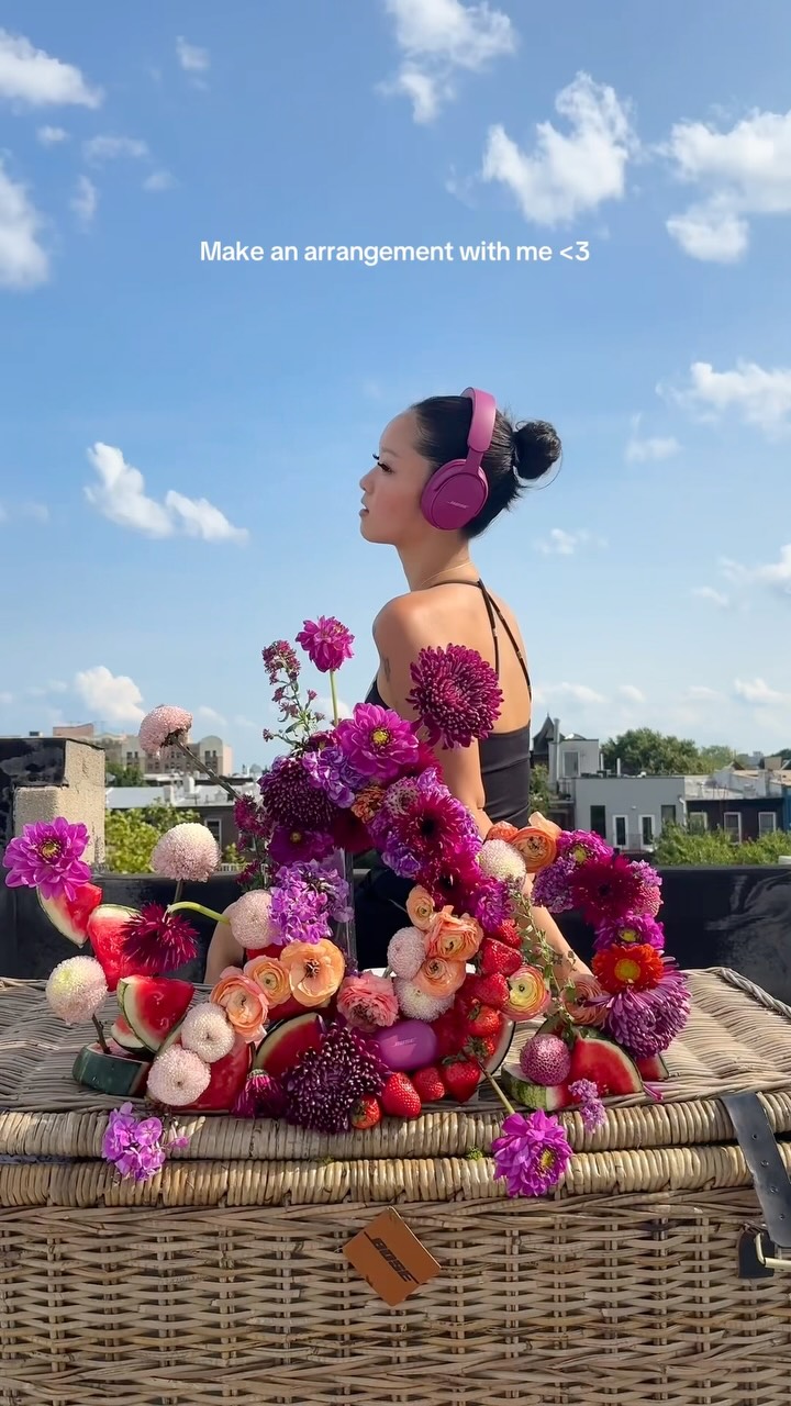 A woman wearing pink headphones is sitting outdoors with a large, colorful arrangement of flowers and fruit in front of her. The text "Make an arrangement with me <3" is overlaid on the image. The background is a bright blue sky with scattered clouds. The woman is looking off to the side, and the arrangement includes various types of flowers, strawberries, and watermelon slices.