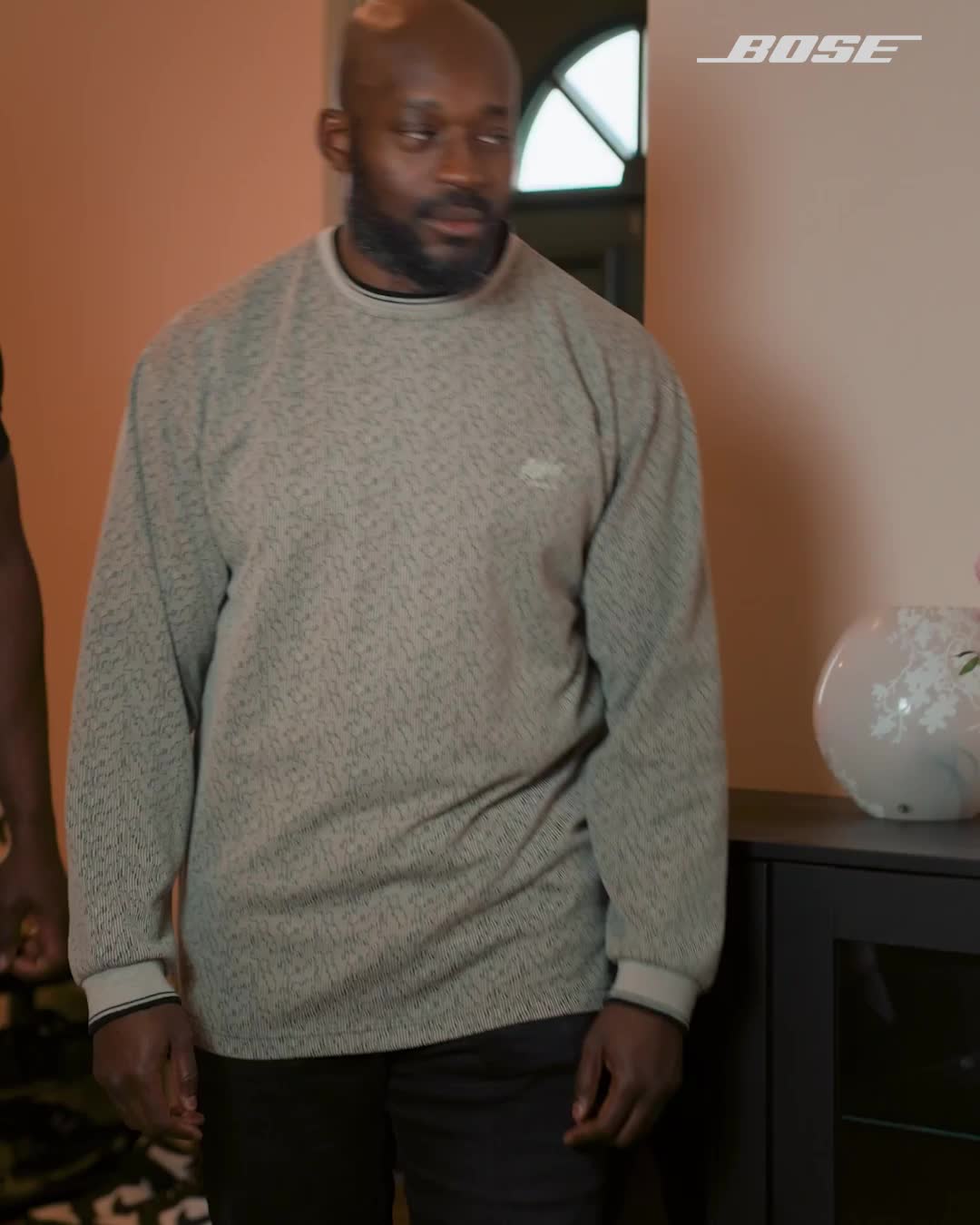 A man with a beard and a bald head is walking forward, wearing a grey patterned sweater and black pants. The Bose logo is visible in the top right corner of the image. The text overlay states, "With 360-degree sound, every scene feels real." The scene appears to be indoors, with a peach-colored wall and a dark cabinet on the right side.